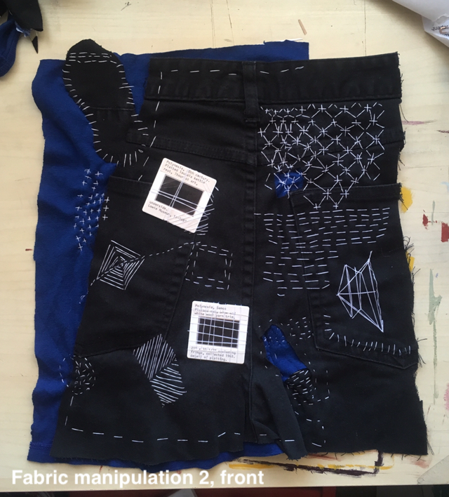 a panel of fabric, mostly the top of some black jeans, with blue fabric attached. it's embroidered and sewn in patterns with white thread. two small film slides are sewn onto it. it's labeled 'fabric manipulation 2, front'