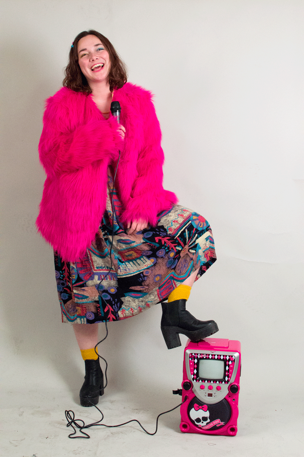 a picture of violet wearing a skirt, yellow socks, platform boots, and a bright pink faux fur coat. she's smiling and holding a microphone and one foot is resting on a monster high-branded karaoke machine.