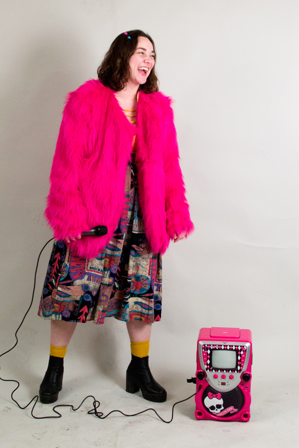 a picture of violet wearing a skirt, yellow socks, platform boots, and a bright pink faux fur coat. she's smiling and holding a microphone, looking off to the side. beside her is a monster high-branded karaoke machine.